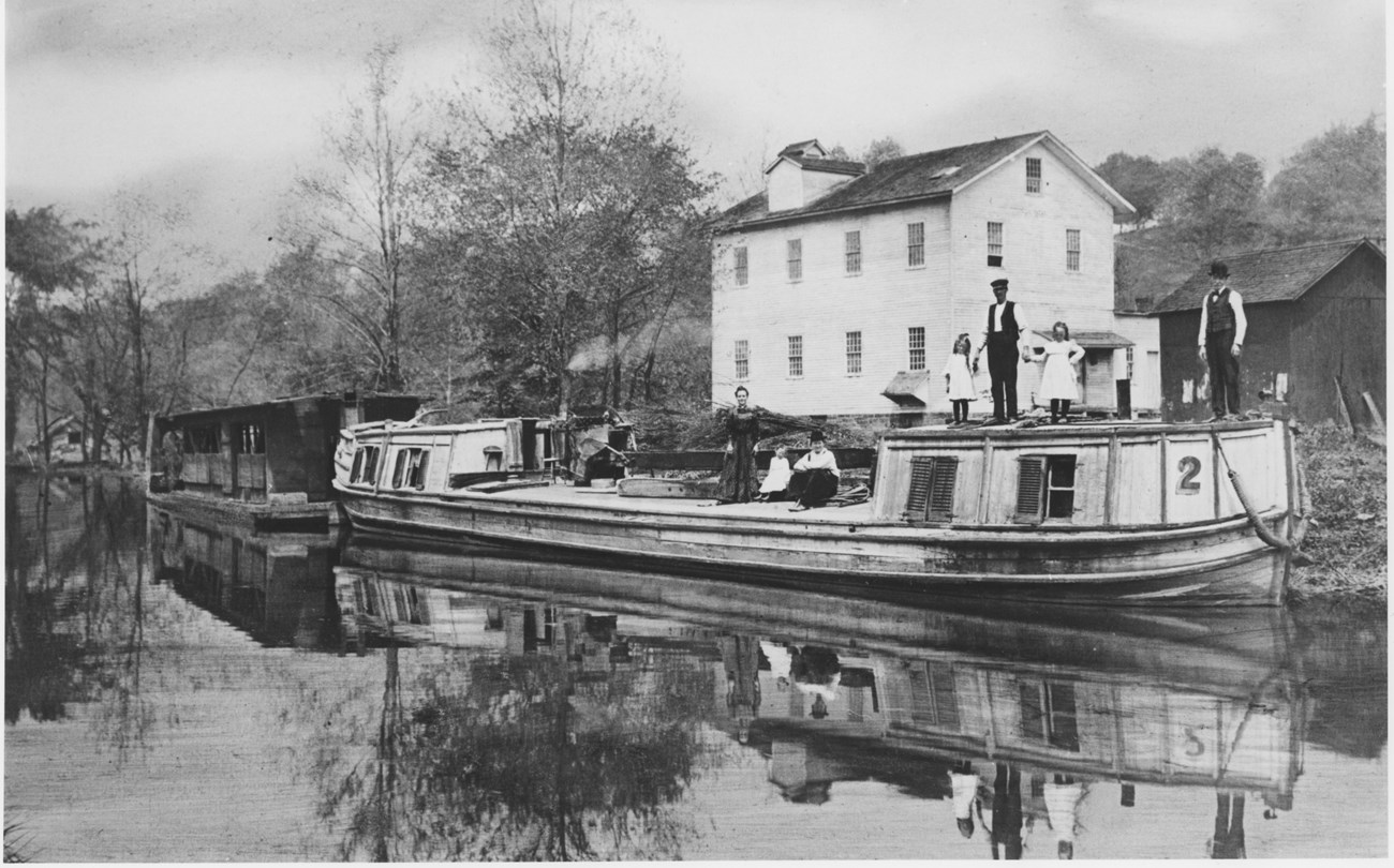 Black and white photo of a group of people – adults and children – standing and sitting on a white canal boat on the water; a white two-story building in the background.