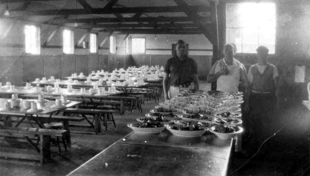 three men stand behind a table set with dishes in a mess hall