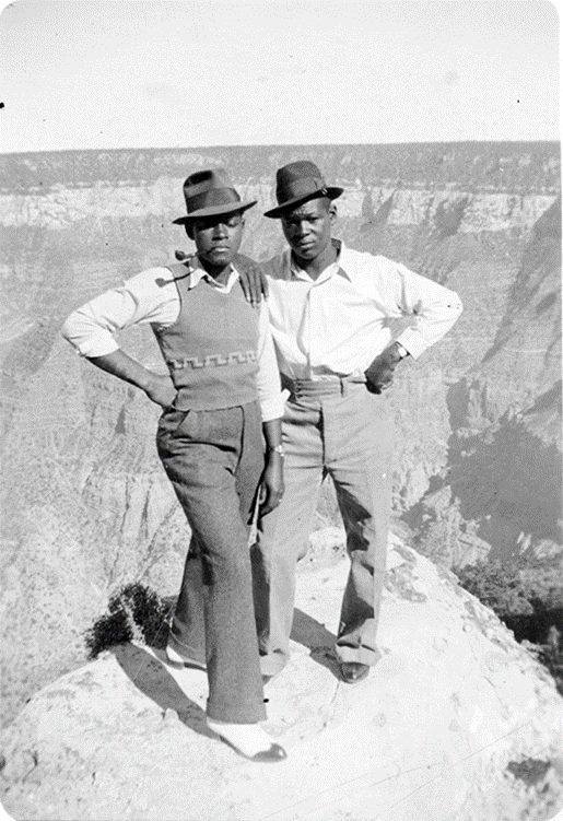 Two Black men stand on the edge of Grand Canyon in nice pants and fedoras.