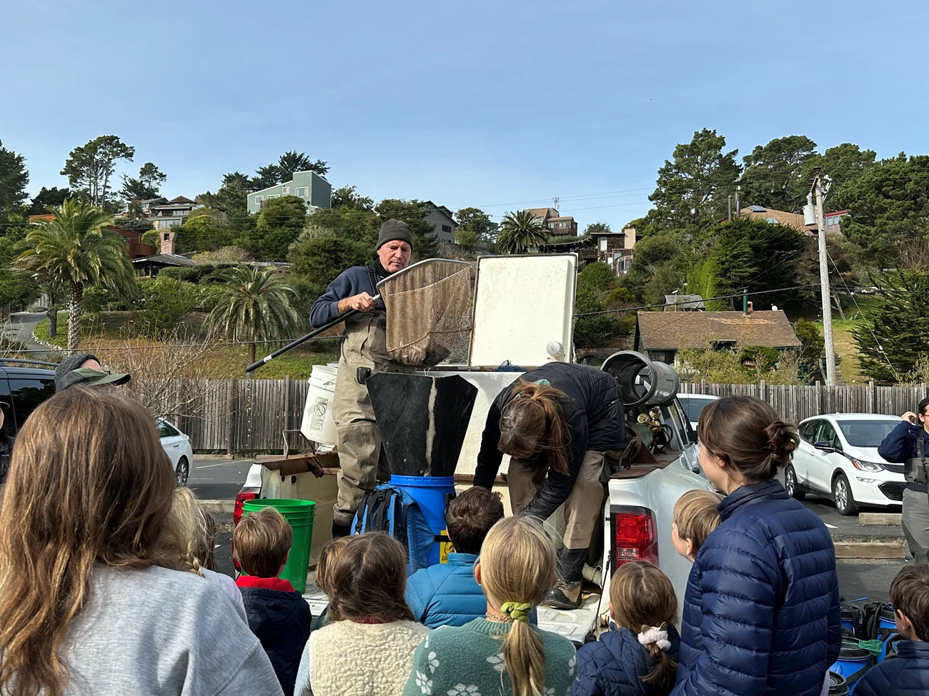 CDFW offloading fish juvenile release People stand on the back of a pickup truck, using a large net and funnel to transfer fish to a blue bucket-backpack in front of a crowd of children and adults.