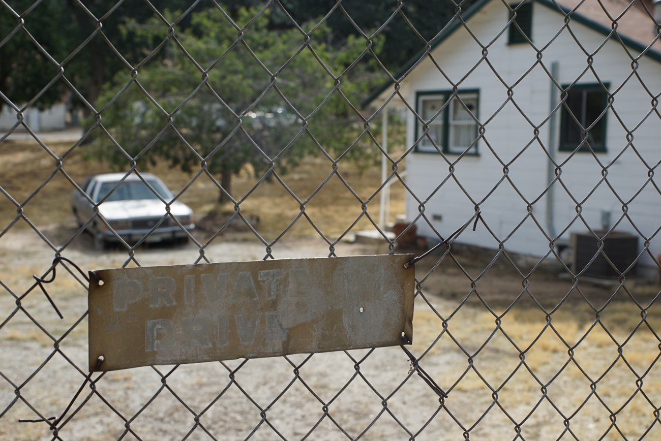Simple white house and dirt driveway seen through chain-link fence. Faded metal sign reads “Private Drive.”