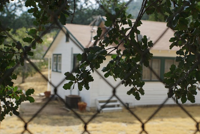 Simple single-story white house with green window trim seen through chain-link fence.