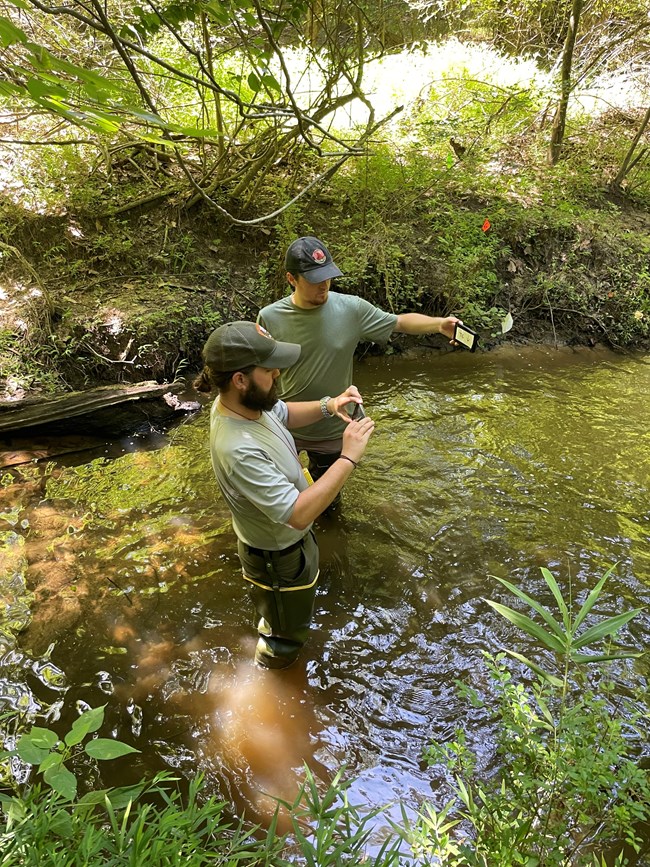 Man holding a phone taking a picture with another man holding a card in the photo. Both are standing in a stream.