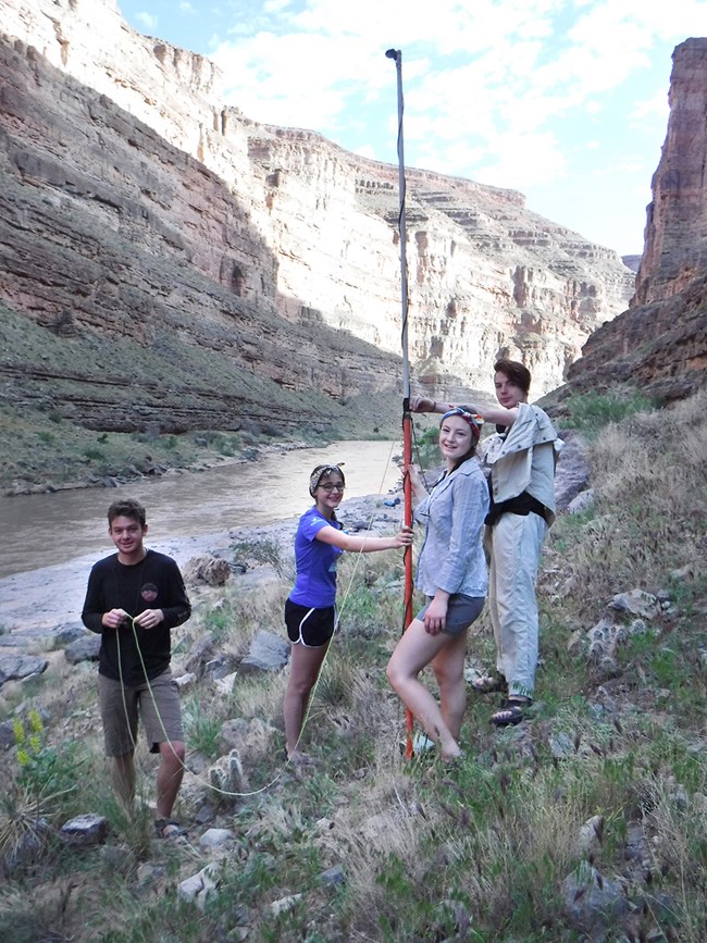 Four students assembling a bat detector uphill from a river. They raise a pole that has a small microphone on top.