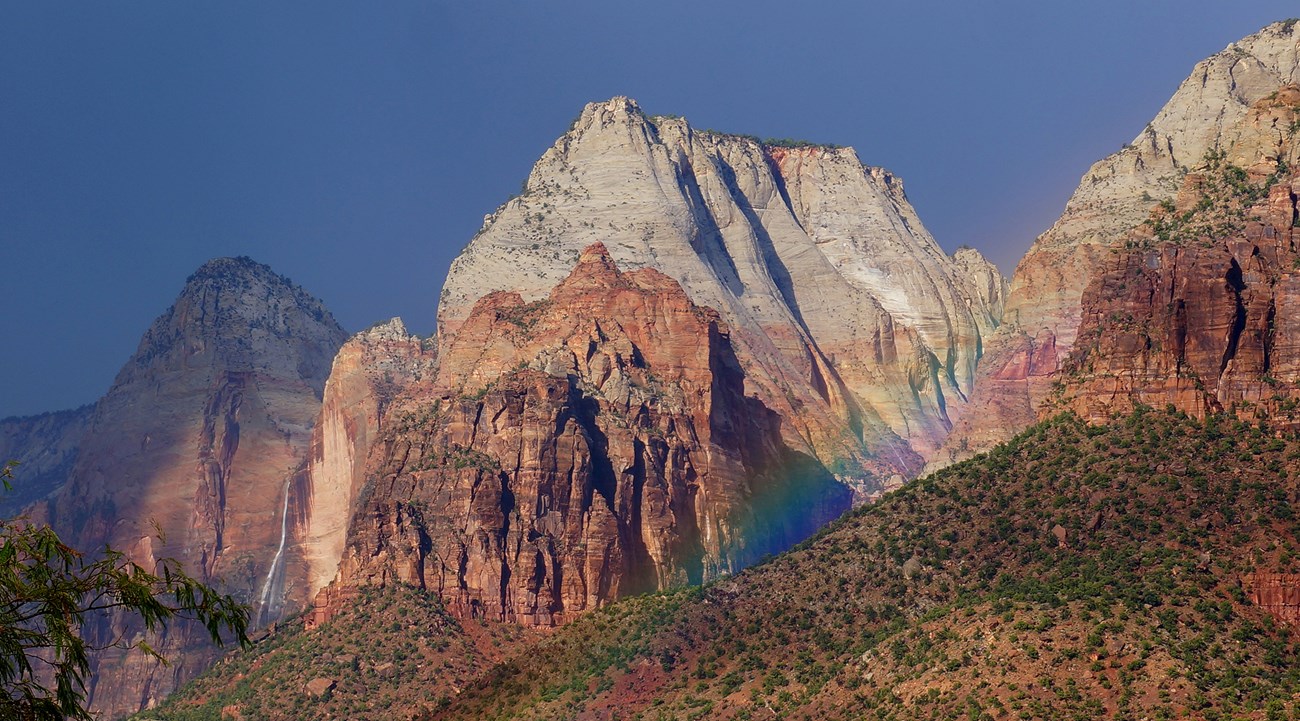 Sunlit sandstone peaks rise under a dark sky as a faint rainbow arcs across the canyon slope.