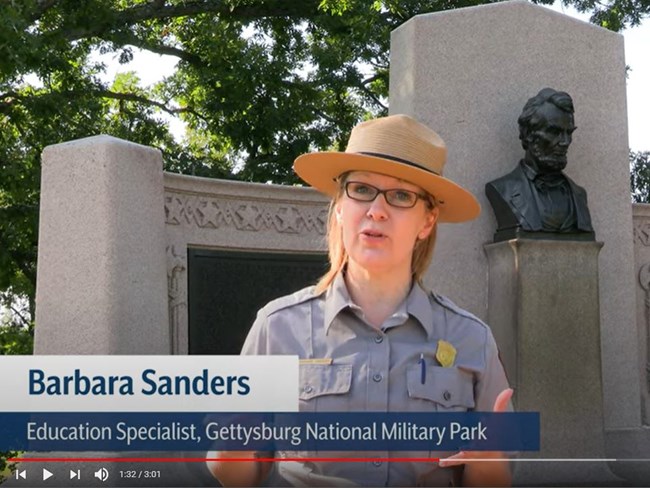 Video screenshot of woman in flathat & park ranger uniform standing in front of memorial