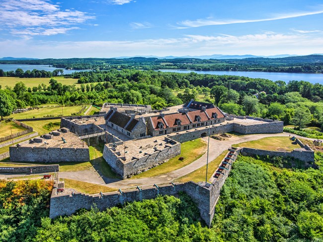 Star-shaped stone fort atop wooded hill, surrounded by lush forest and overlooking body of water.