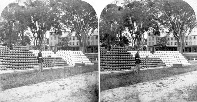 Black and white stereograph of pyramid-like piles of cannons on a lawn with a building in the background.