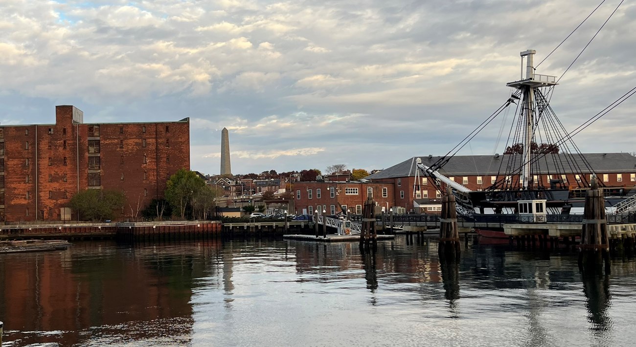 view from the water of the Charlestown Navy Yard, with the brick hoosac warehouse building to the left and the USS Constitution to the right