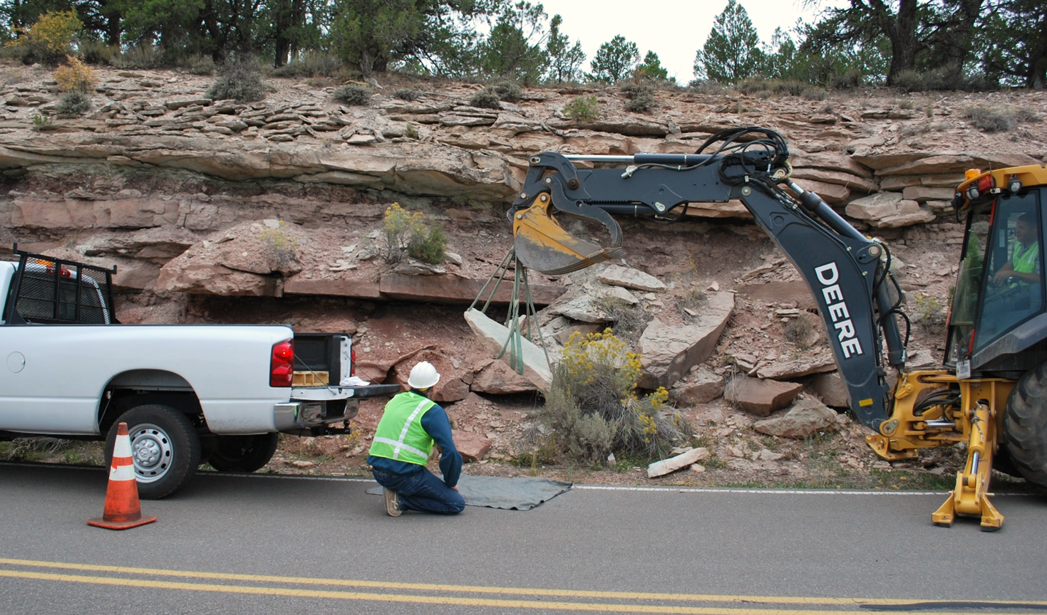 Pickup truck and backhoe with workers move boulder at the base of a roadside bluff.