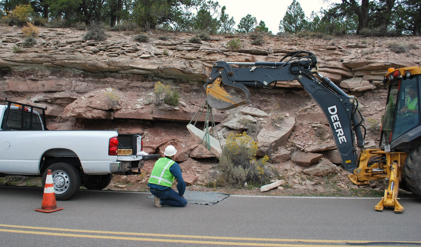 COLM Figure 1 Pickup truck and backhoe with workers move boulder at the base of a roadside bluff.