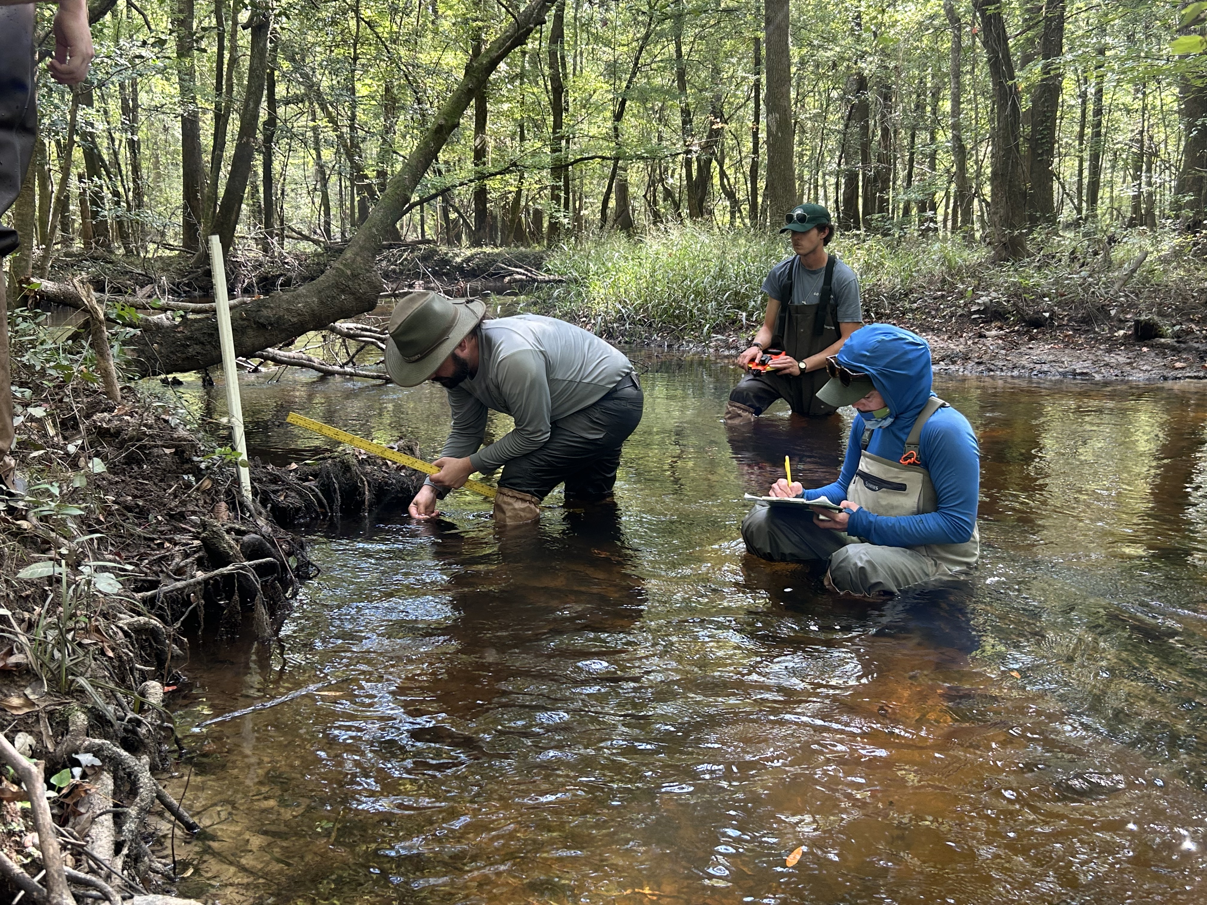 Man kneels over to look a soil standing in a stream. Woman with waders sits in the stream writing on a clipboard and another man holds a tape