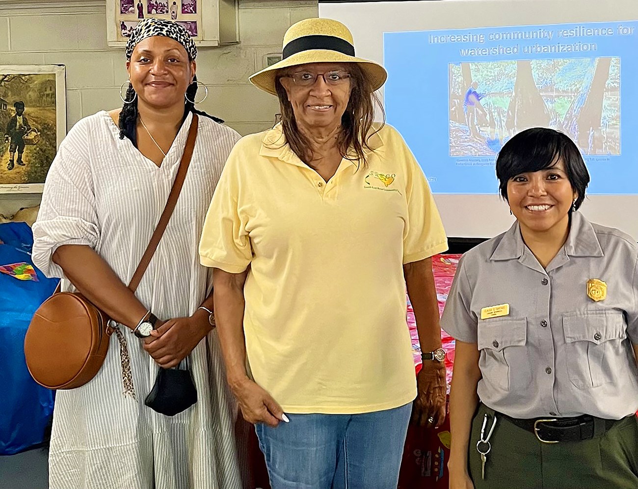 Three women, one of whom wears a National Park Service uniform, stand smiling in front of a projector screen. The slide on the screen reads "Increasing community relilience for watershed urbanization."