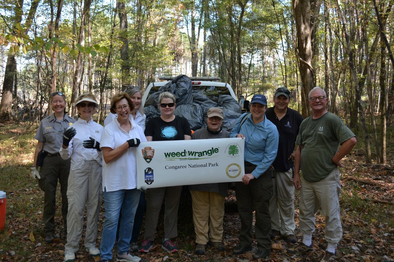 Southeast Coast Invasive Plant Management Team nine adults hold a banner that says Weed Wrangle Congaree National Park in front of pickup truck filled with garbage bags.