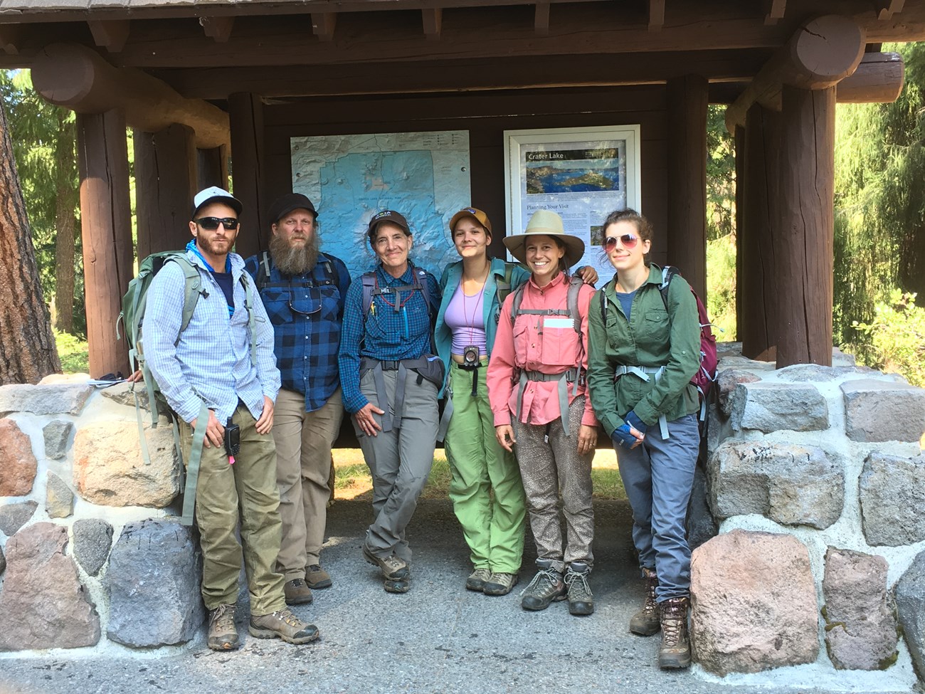 Six people wearing backpacks and hiking boots pose for a photo before an outdoor map of Crater Lake.