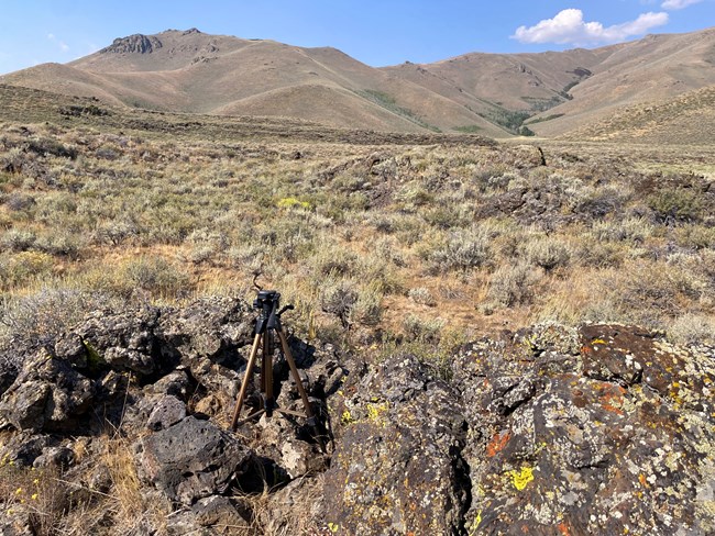A trail camera sits on a tripod in a rocky lava outcrop at Pronghorn Pass. Sagebrush vegetation dominates the rocky pass and the pioneer mountains protrude out of the landscape in the nearby distance.