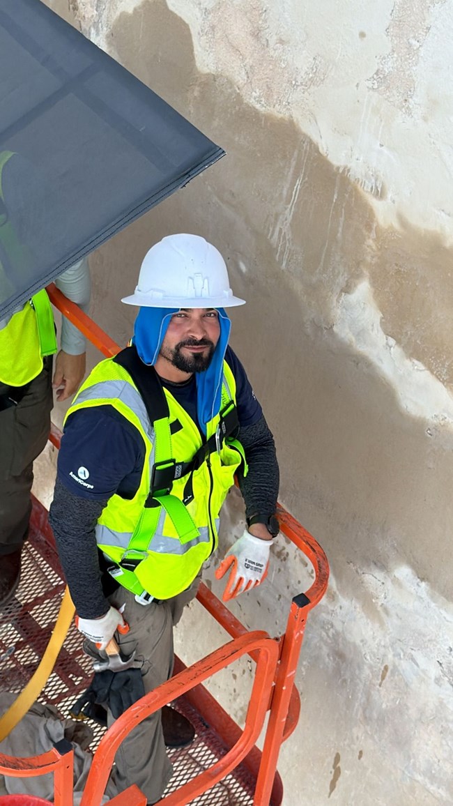 Worker stands on orange aerial boom lift next to La Fortaleza wall at San Juan National Historic Site wearing blue towel under helmet, white helmet, neon green safety vest, and gloves with hammer in hand.