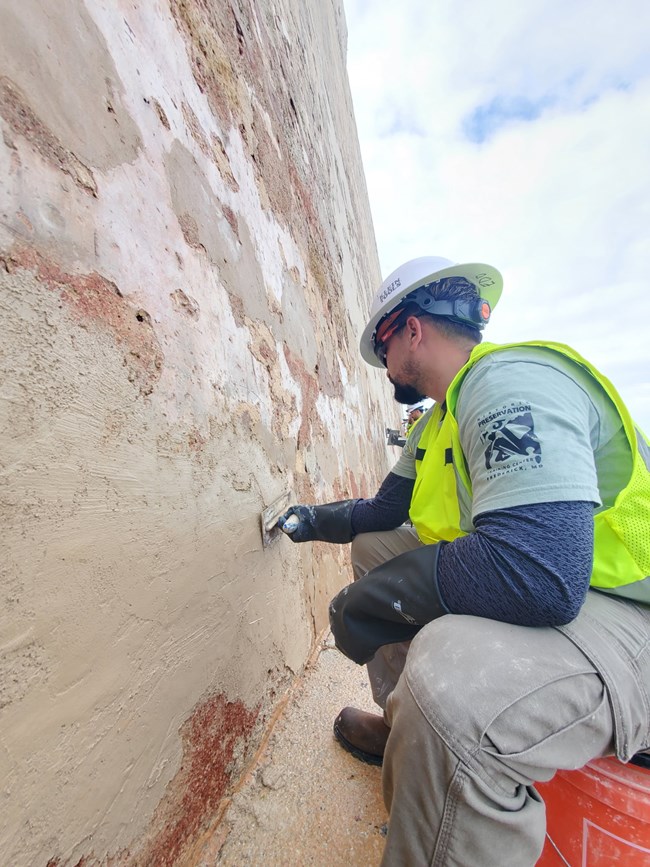 Worker spreading beige cement on beige fort wall. Worker wears white helmet, black sunglasses, neon green safety vest, t-shirt, black gloves, and green cargo pants. He sits on an orange bucket.