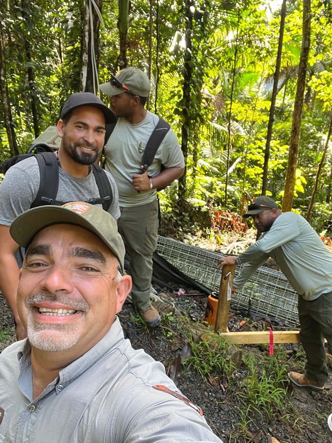 Five men capture selfie in forest setting at El Yunque National Forest. Trees surround the men, all wearing grey shirts and green hats with two of the men in backpacks.