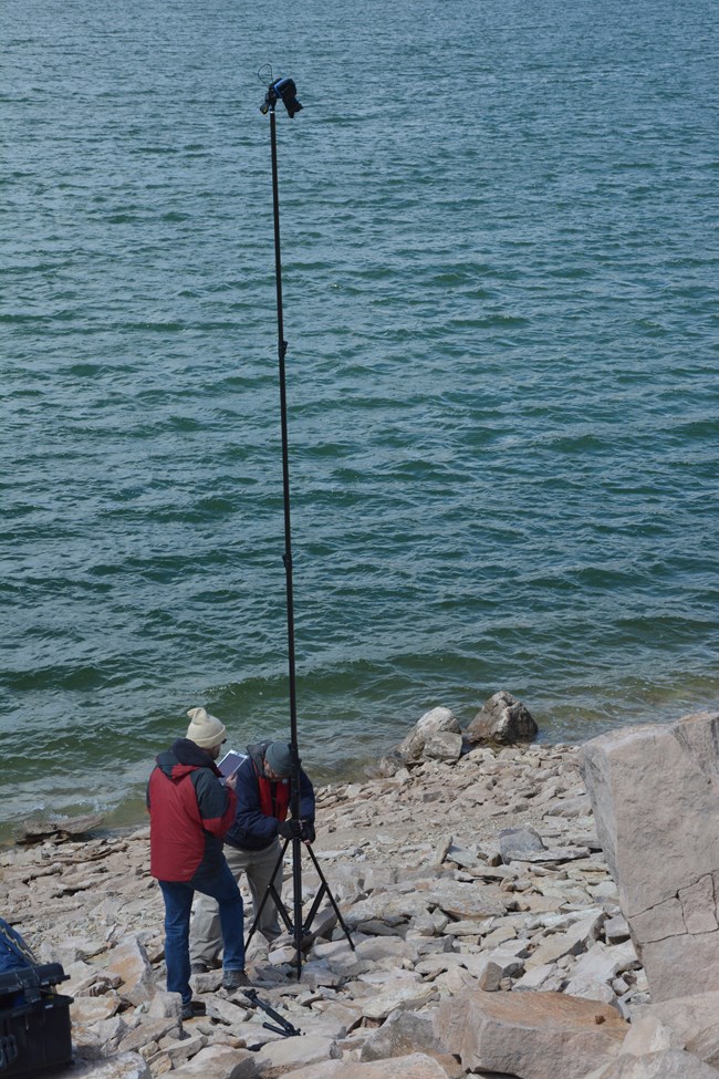 two people with a tall camera pole standing near a lake shore