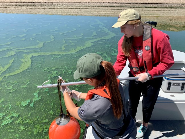 two women on a boat fish a buoy out of green algae ridden water