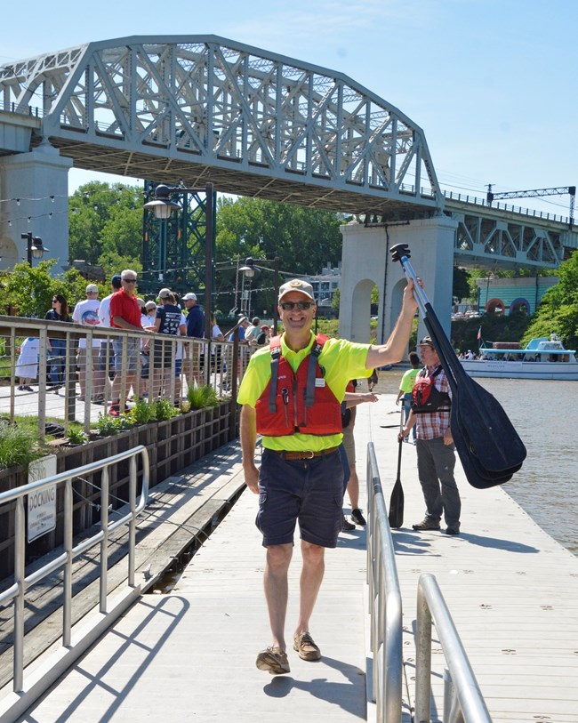 A man wearing a life jacket walks along a dock carrying canoe paddles