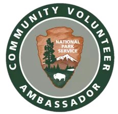 Circular badge with a green outer perimeter displaying the words 'Community Volunteer Ambassador' in white capital letters. At the center, the National Park Service emblem features an arrowhead shape with a Sequoia tree, mountain, and bison, along with th