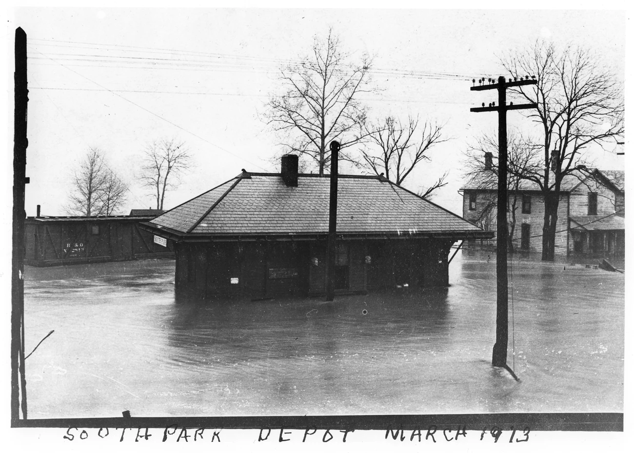 CVNP_Flood_Brecksville Black and white flood scene showing a one-story station, a train car, an electric pole, and a two-story wooden house. The depot has a “South Park” sign.