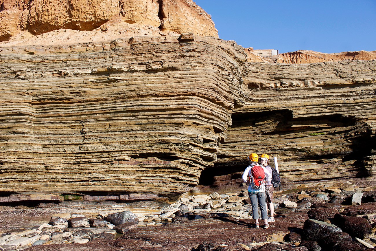 two people waring helmets stand next to a rock formation with distinctive layering