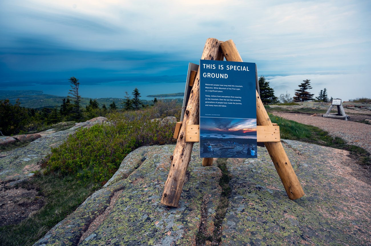 Wood structure with three legs and attached display panels placed on a granite boulder on a mountain summit.
