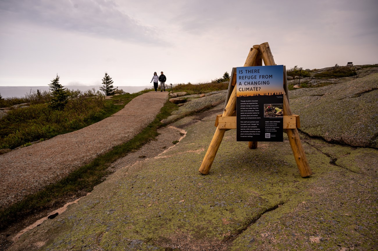 Wood structure with three legs and attached display panels placed beside a stone pathway on a mountain summit. A woman and man walk together along the path. Wood structure with three legs and attached display panels placed beside a stone pathway on a mountain summit. A woman and man walk together along the path.
