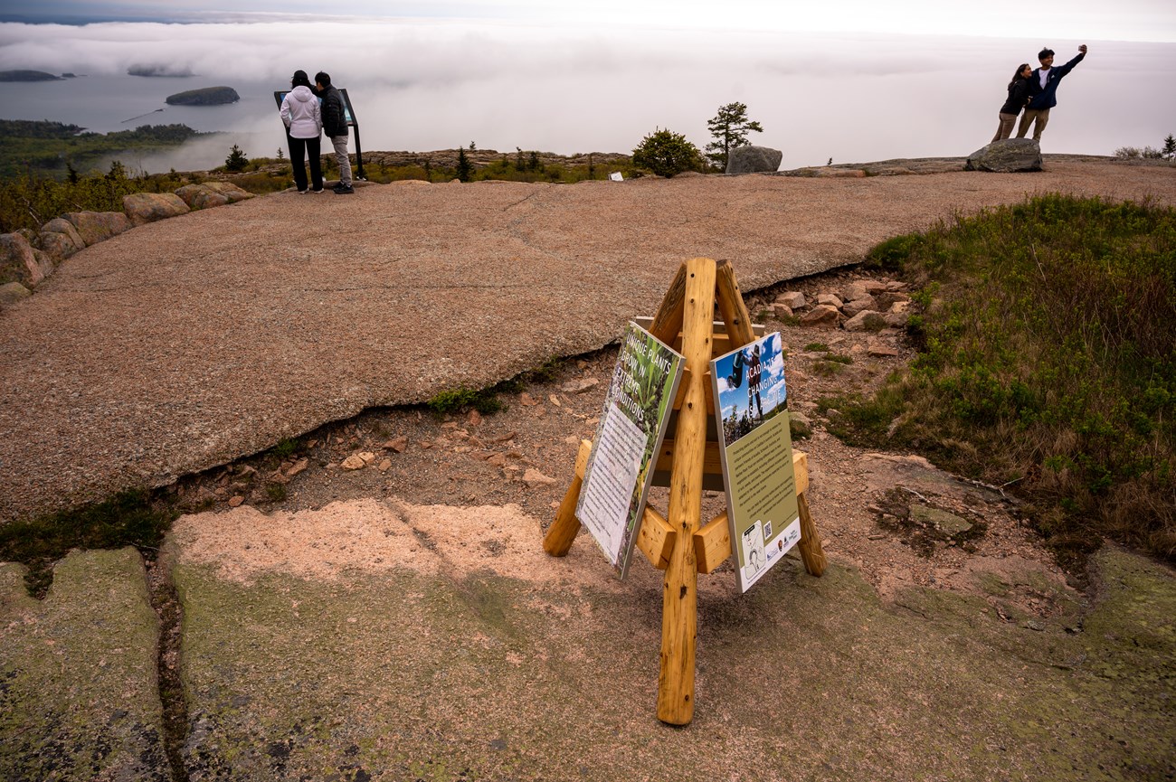 Wood structure with three legs and attached display panels placed beside a stone pathway on a mountain summit. Visitors look at a wayside and snap a selfie. Wood structure with three legs and attached display panels placed beside a stone pathway on a mountain summit. Visitors look at a wayside and snap a selfie.