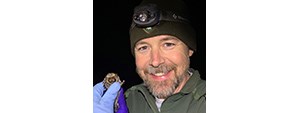 Allen in NPS green jacket and hat, wearing a headlamp, smiling for the camera and hollding up a bat with purple rubber gloved hands.