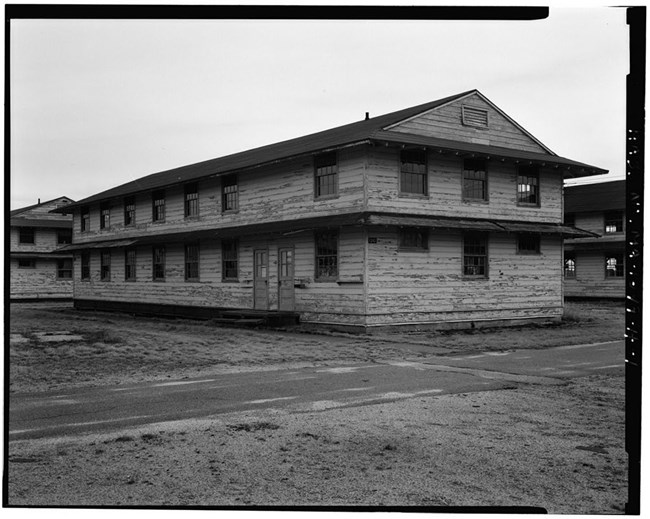 Perspective view of south side (front) and east side of two-story barracks building covered in wood paneling with worn and chipped paint