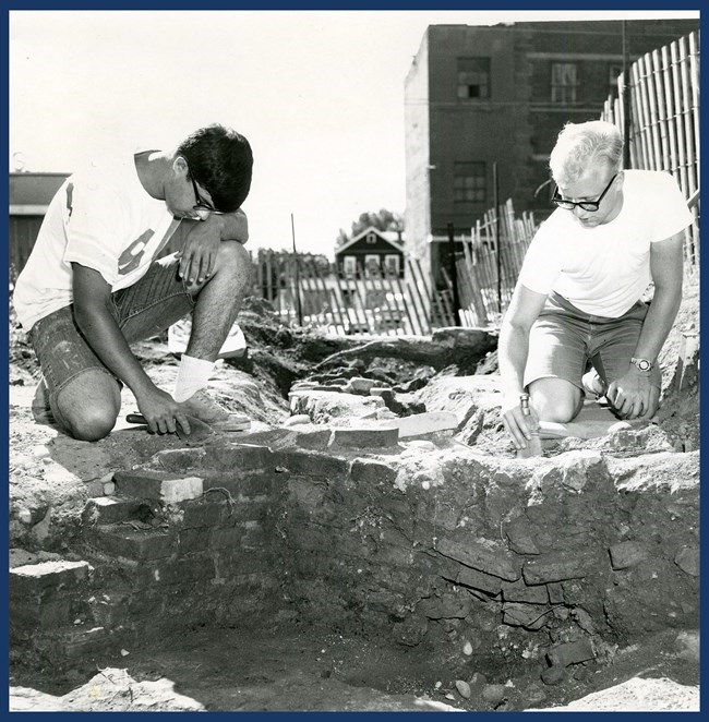 An archeological excavation. 2 men sit above a pit with a brick foundation exposed. They hold trowels.
