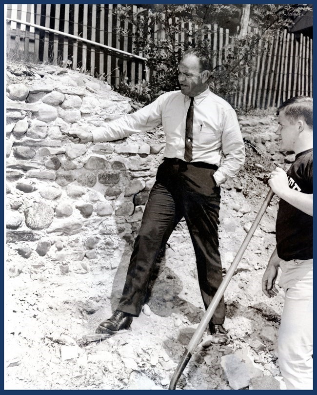 A man in slacks, shirt, and tie points to a rocky wall.