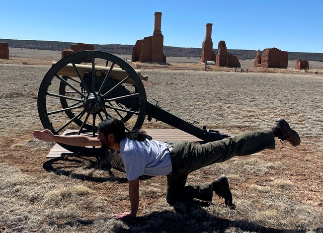 Park Ranger kneeling on right knee with left leg extended backward and right arm extended forward with cannon and adobe buildings in background