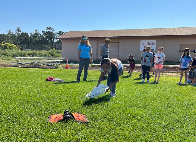 A girl bends over holding an insect net to capture a monarch puppet in green grass.