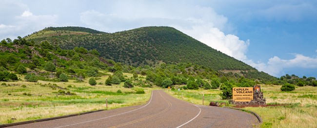 Established in 1916, Capulin is the oldest of the High Plains Group of historic sites. Entrance to Capulin Volcano National Monument with sign and cinder cone volcano covered with green vegetation under blue sky