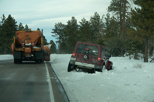 A large truck drives down a road past a maroon jeep stuck in snow just off the pavement.