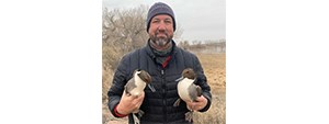 Man in a hat and coat holding a duck under each arm.