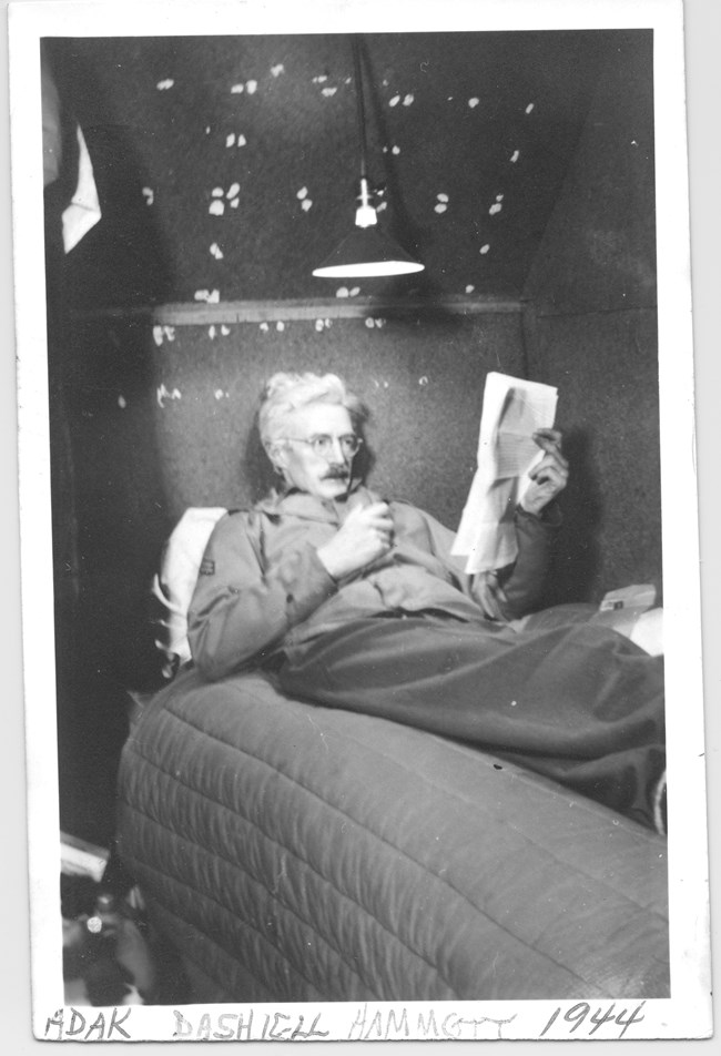 Gray-haired man lying on a bed reading a newspaper.