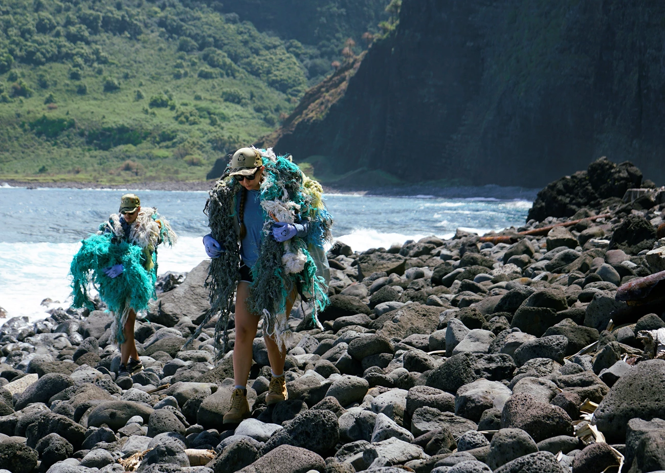 Carrying out marine debris two people carry large bundles of collected marine fishing nets and other marine debris along a rocky shoreline with steep mountain cliffs in the background