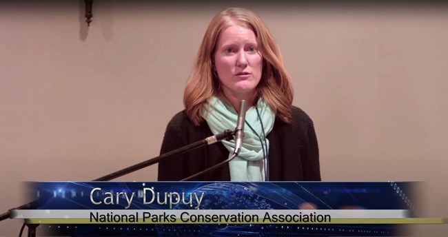 The presenter standing at a lectern. Watch a non-audio described version of this presentation on YouTube.
