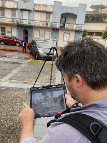 A man looking at an iPad. The iPad is showing the viewer an image of a historic building. In the background is a tripod with a laser scanner facing a road.  Cars are parked on the road with a cream and light blue older building across the street.