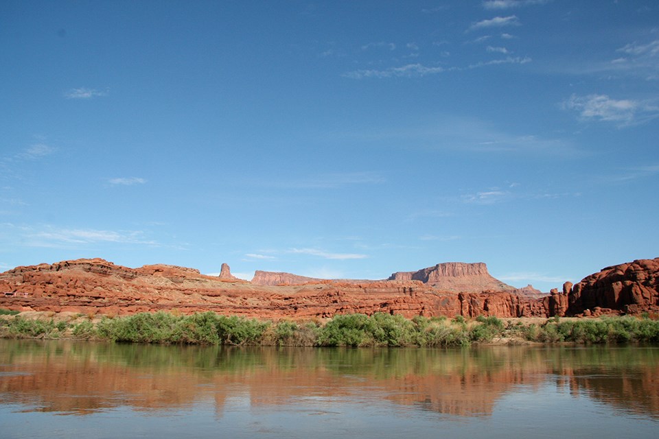 Body of water in the foreground reflects the green shrubs on the bank, red rock formations in the background and a brilliant blue sky with wispy clouds.