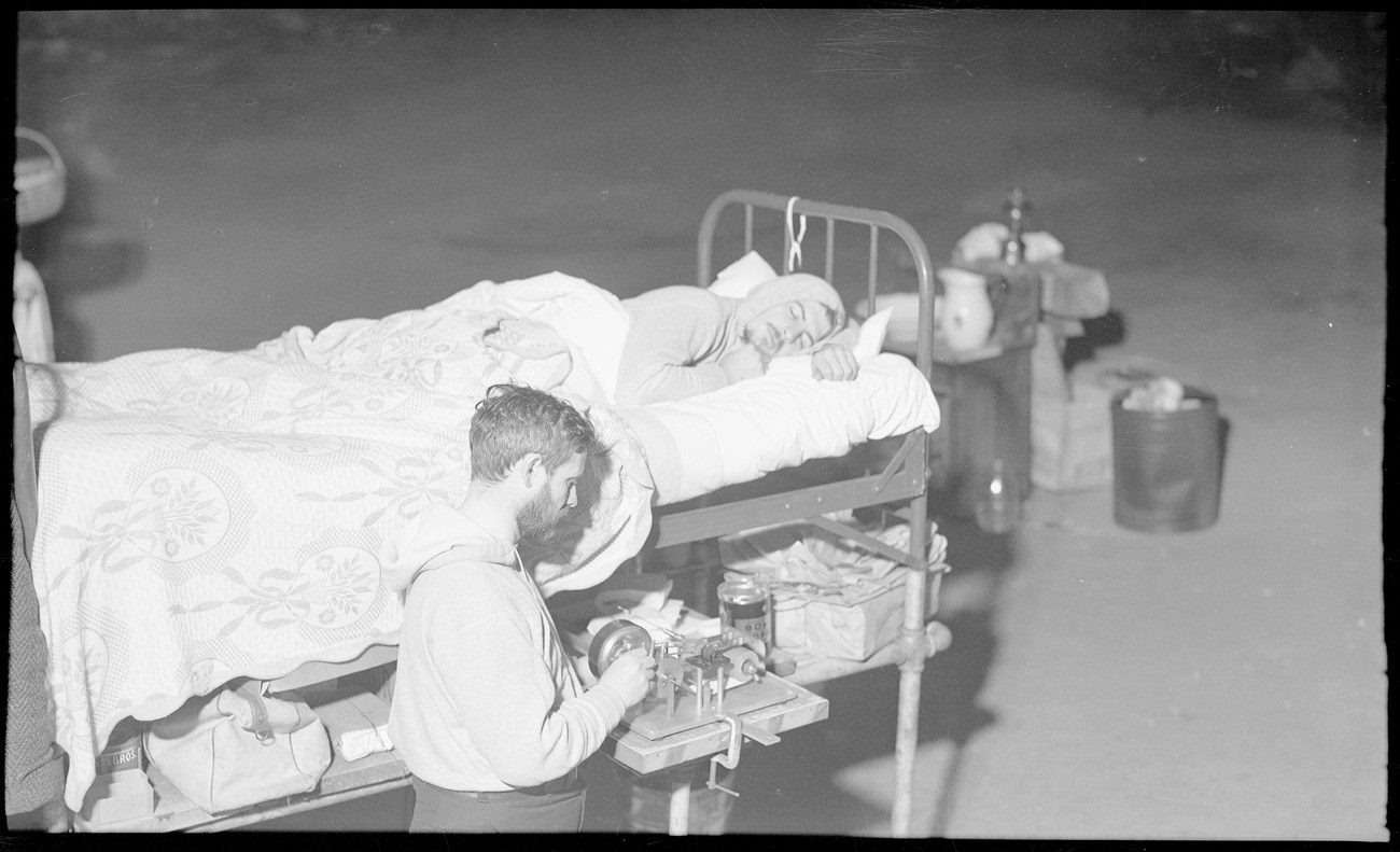 A man uses a piece of equipment to take data readings while another man sleeps in a metal framed bed.