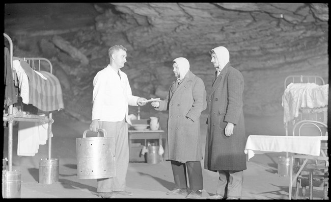 Three men stand in a cave passageway near assorted metal-framed furniture. A man wearing a waitstaff uniform holds a metal food container and gives an envelope to one of the other men.