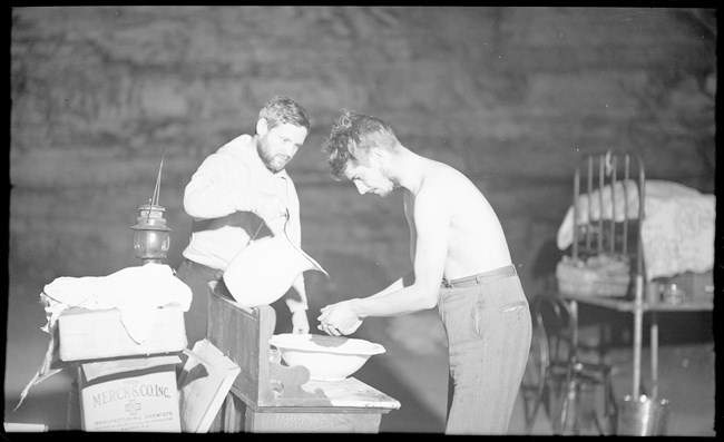 Two men stand near a wash stand in an underground cave passage. One washes his hands while the other man pours water from a large pitcher.