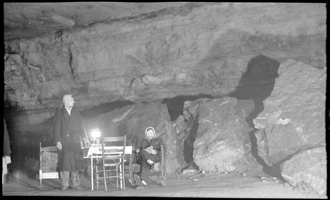 Two men in an underground cave passageway, one stands while the other reads by lanternlight while seated at a table.